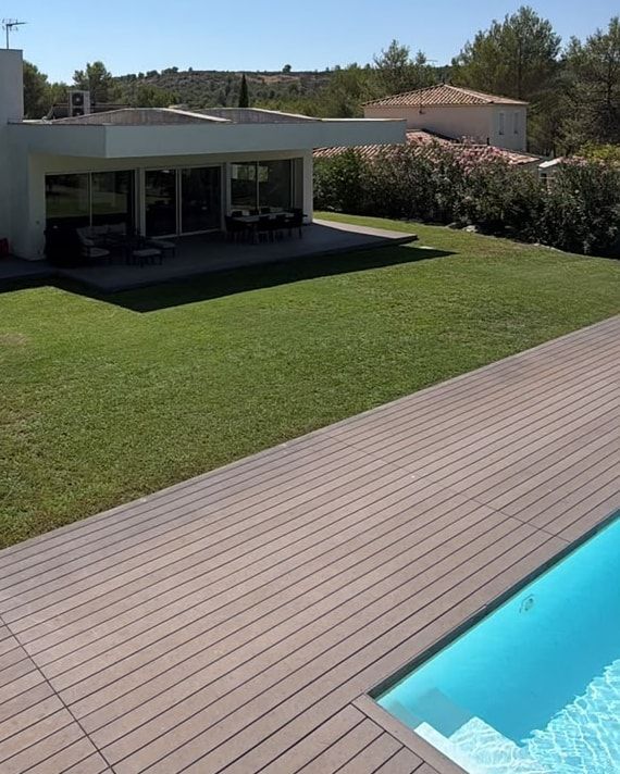 Plage de piscine en lames composite gris-brun, pelouse et villa cubique blanche entre les pins, à Grabels