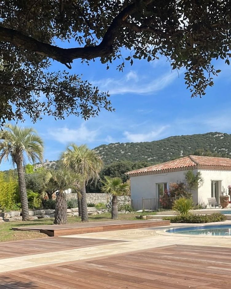 Terrasse en ipé et dallage clair au bord d’un bassin, palmiers, maison aux tuiles et colline de garrigue, à Saint-Gély-du-Fesc