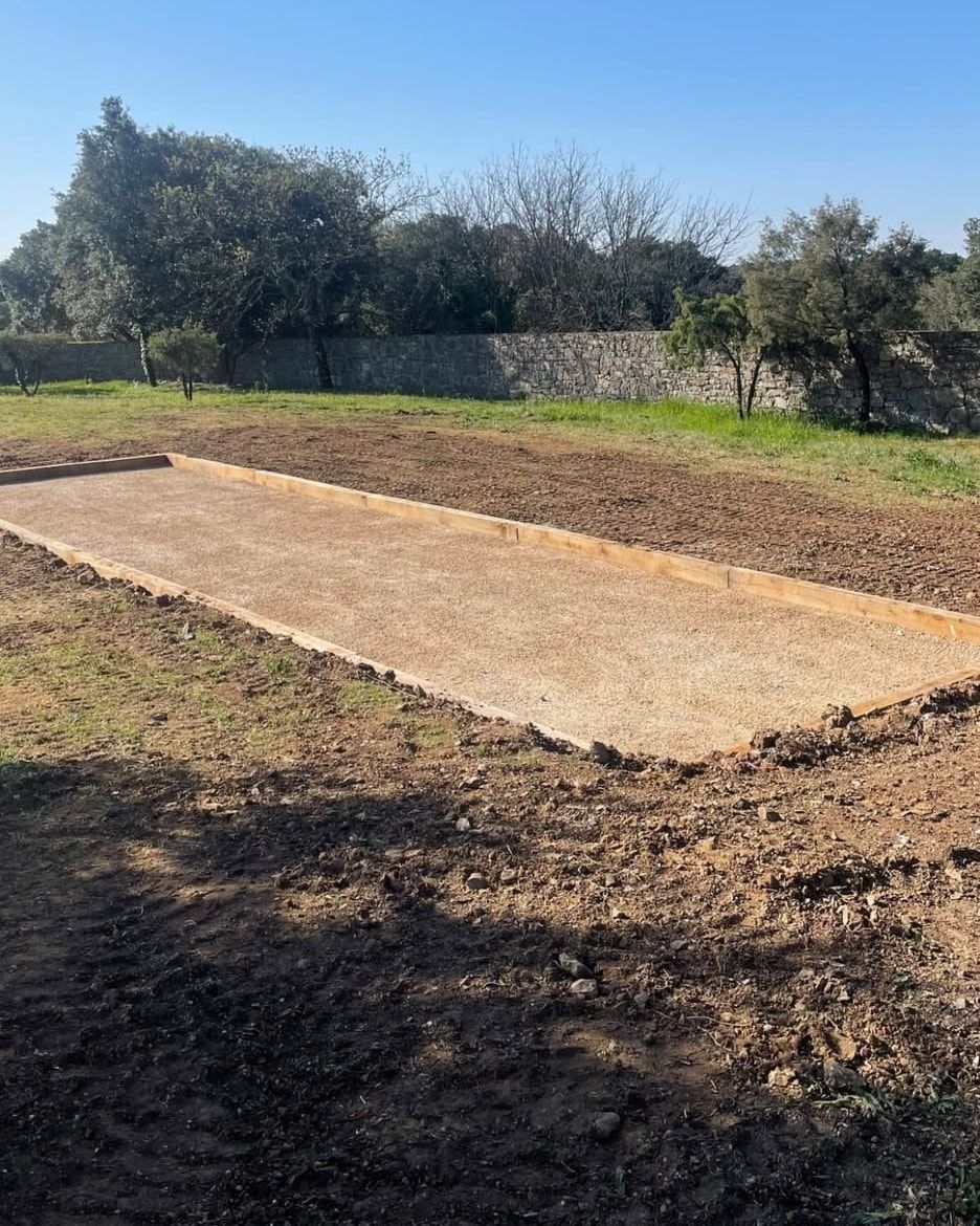 Terrain de pétanque bordé de bois, gravillon stabilisé, mur en pierre sèche et chênes verts