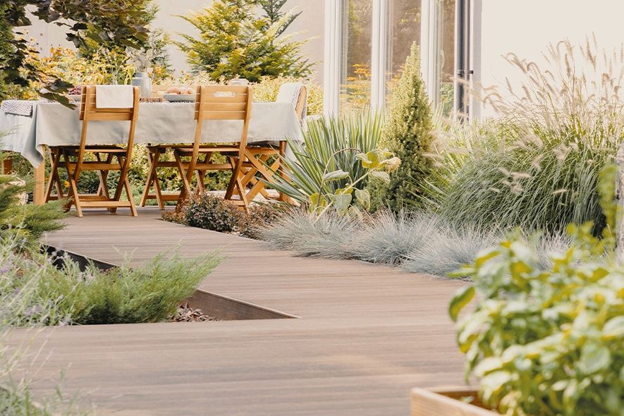 Terrasse en lames de bois composite bordée de graminées, table et chaises devant une baie vitrée